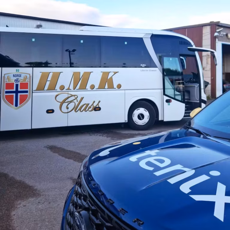 White 'H.M.K. Class' tour bus with Norwegian flag emblem parked near a blue Saga Tenix vehicle, highlighting fleet control and maintenance solutions for electric vehicles.