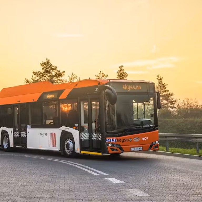 Modern public bus with an orange and white "Skyss" design, driving on a curved road at sunset with trees in the background