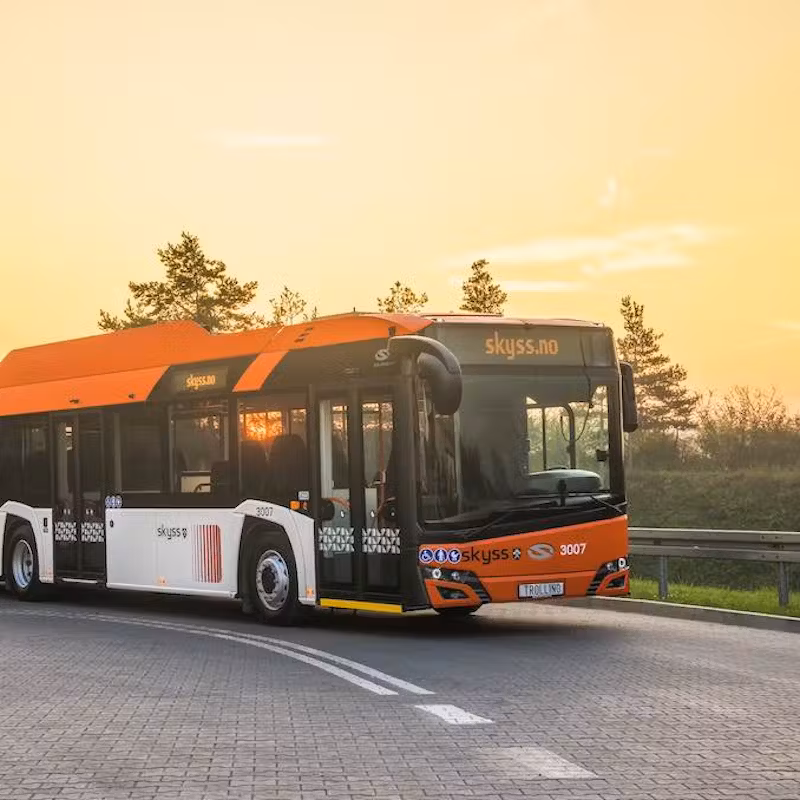 Modern public bus with an orange and white "Skyss" design, driving on a curved road at sunset with trees in the background