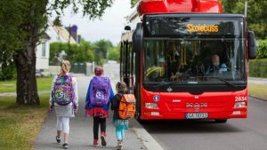 Three children with colorful backpacks walking towards a red MAN school bus ('Skolebuss') parked on a suburban street with the driver visible inside.