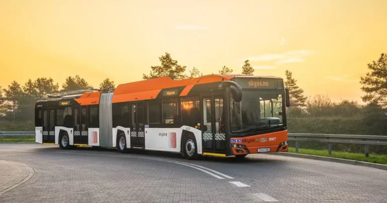 Articulated public bus with an orange and white design, branded with "Skyss" and "skyss.no," driving on a curved road at sunset with trees in the background