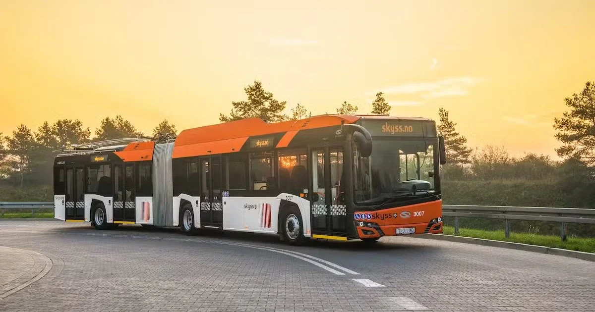 Articulated public bus with an orange and white design, branded with "Skyss" and "skyss.no," driving on a curved road at sunset with trees in the background