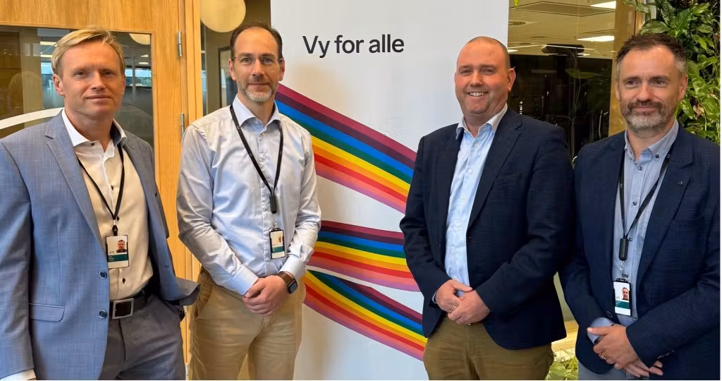 CEO Torbjørn Krøvel - and representativs for VY in business attire wearing ID badges, standing in front of a 'Vy for alle' banner with a colorful rainbow design in an office setting.