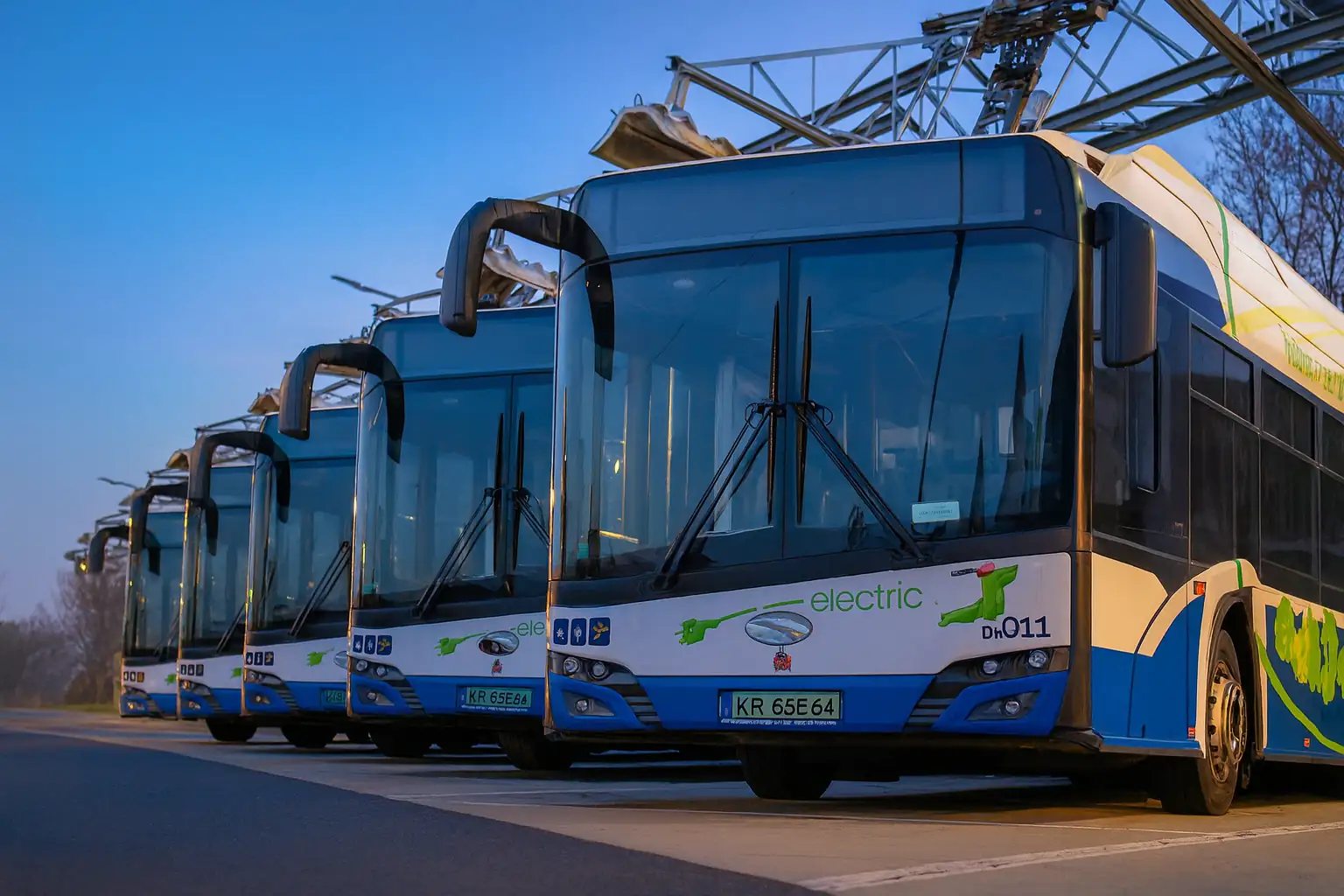 Tenix Charge Operations for Modern Electric Fleets Line of blue and white electric buses parked at a depot with overhead charging equipment, ready for service at dawn.