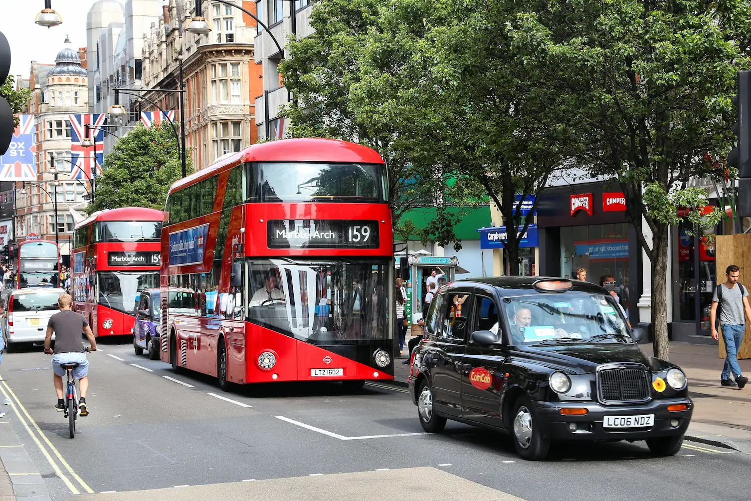 Red double-decker buses and a black taxi navigating a busy London street, representing urban fleet tracking and management challenges.